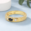 A textured yellow gold ring featuring a central oval black onyx stone and two small round clear stones, displayed on a light blue surface with a white ceramic object in the background.