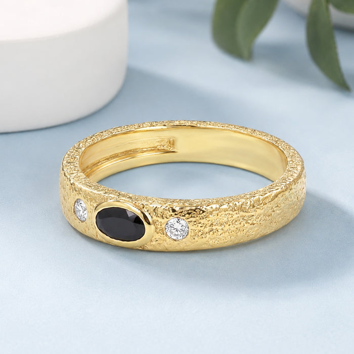 A textured yellow gold ring featuring a central oval black onyx stone and two small round clear stones, displayed on a light blue surface with a white ceramic object in the background.