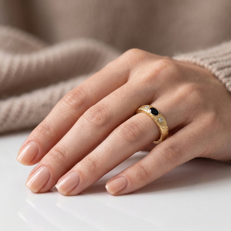 A close-up of a hand wearing a gold ring with a black onyx oval center stone and small clear accent stones, resting on a white surface with a soft beige knit background.