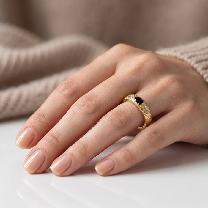 A close-up of a hand wearing a gold ring with a black onyx oval center stone and small clear accent stones, resting on a white surface with a soft beige knit background.