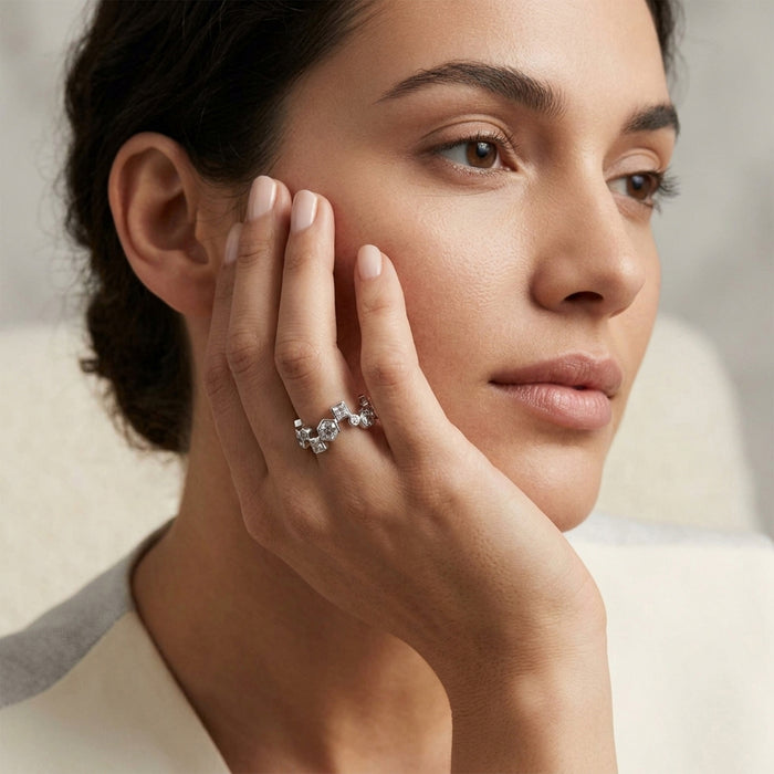 A close-up lifestyle shot of a woman wearing a delicate silver ring with small clear stones on her finger, resting her hand on her cheek against a soft, light beige background.