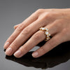 A close-up of a hand wearing a gold ring set with multiple clear gemstones, photographed against a dark reflective surface with a soft gray background.