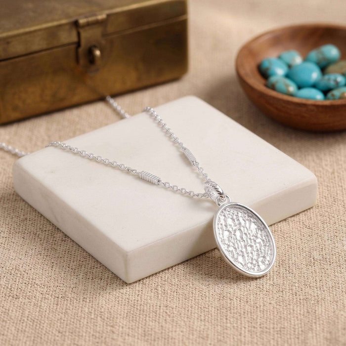 A silver oval pendant necklace with a textured hammered finish, resting on a white stone block. The necklace is styled with a rustic bronze box and a small wooden bowl of turquoise beads on a beige linen background.