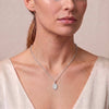 A close-up of a woman wearing a delicate silver chain necklace with a small oval pendant, styled with a cream-colored top against a soft beige background.
