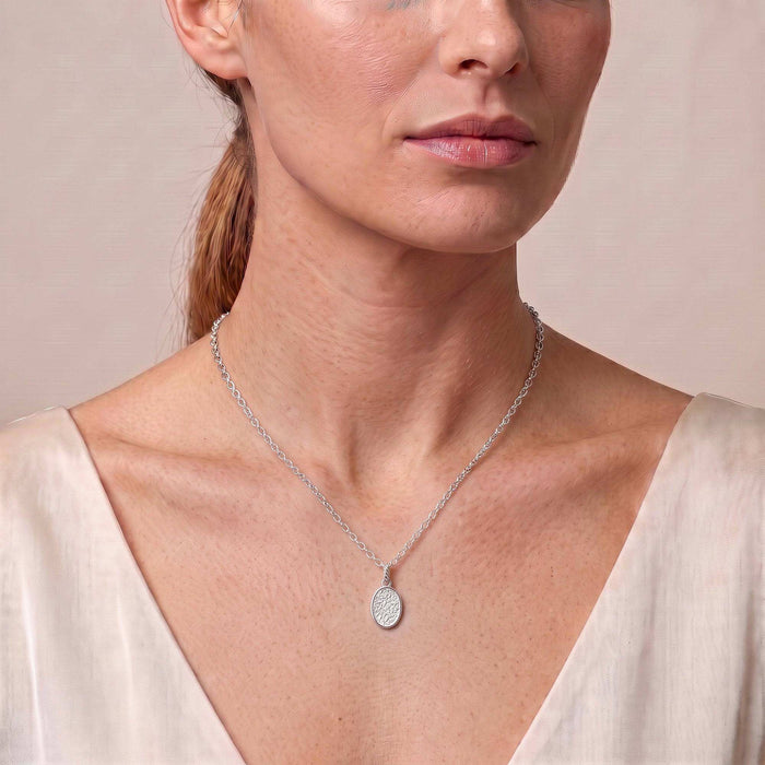 A close-up of a woman wearing a delicate silver chain necklace with a small oval pendant, styled with a cream-colored top against a soft beige background.