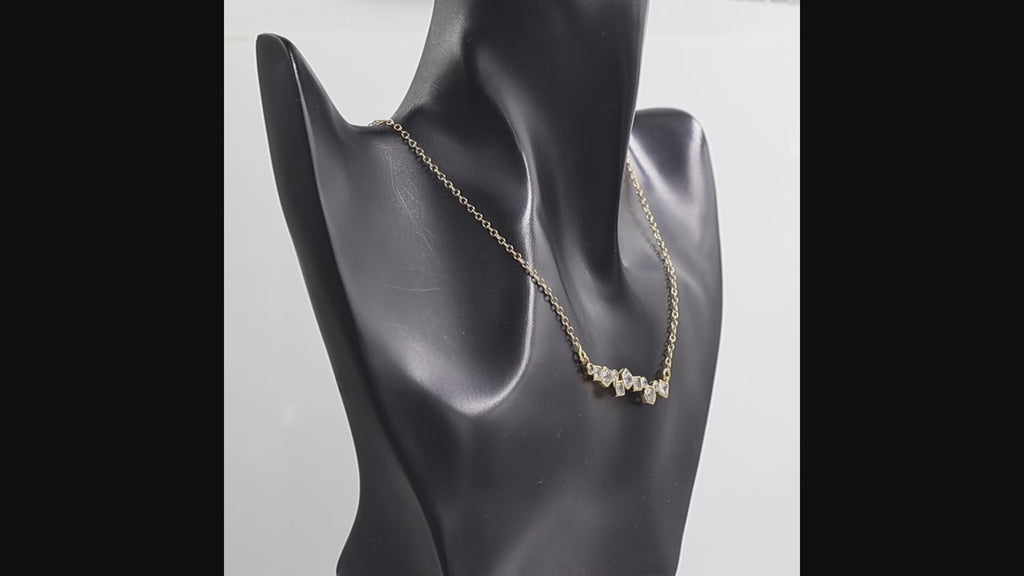 A gold necklace featuring a horizontal bar of clear gemstones in various shapes, including hexagons, squares, and round bezels, suspended from a delicate gold chain against a white background.